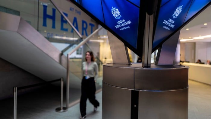 A woman walks past a display screen showing the London Stock Exchange logo.