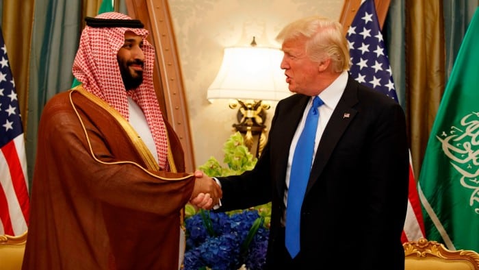 The two men are shaking hands. Saudi and US flags are in the background