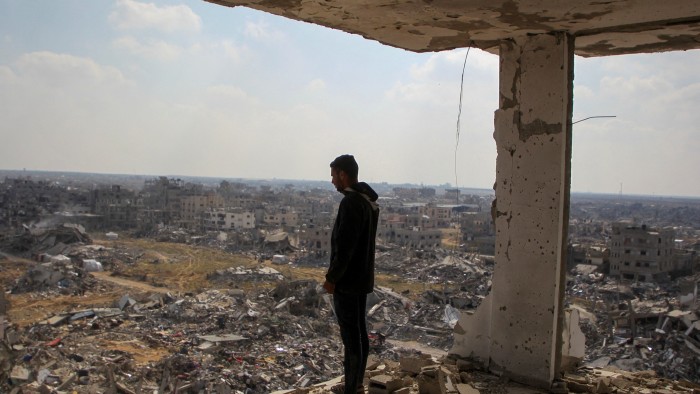 A man stands on the edge of a damaged building, overlooking extensive rubble and destruction in Rafah following an Israeli offensive.