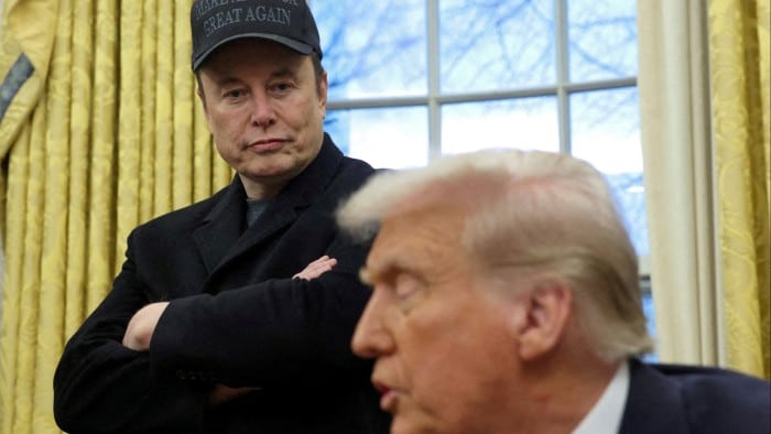 Elon Musk stands with arms crossed, wearing a black jacket and a black Make America Great Again cap, listening as Donald Trump speaks in the Oval Office