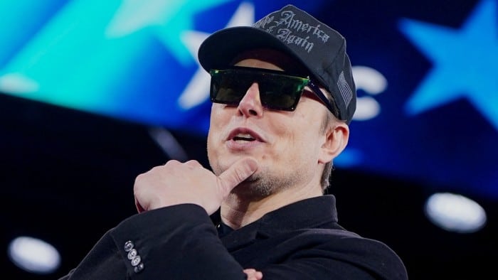 Elon Musk wears sunglasses and a black Make America Great Again cap, while gesturing with his thumb under his chin onstage at the Conservative Political Action Conference