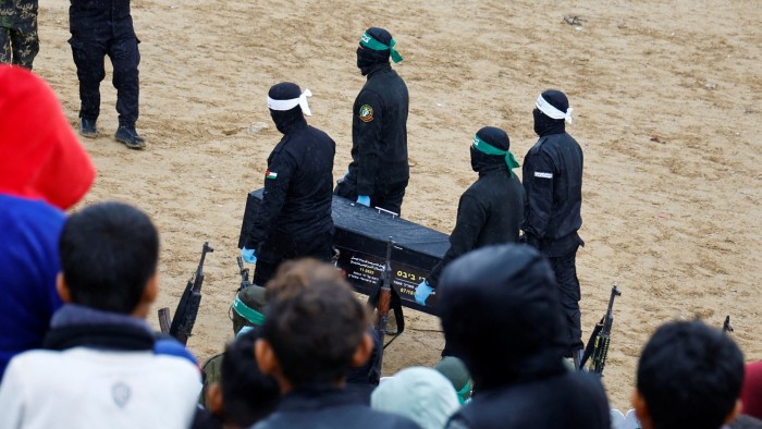 Masked individuals dressed in black with green and white headbands carry a coffin across a sandy area, surrounded by onlookers holding guns