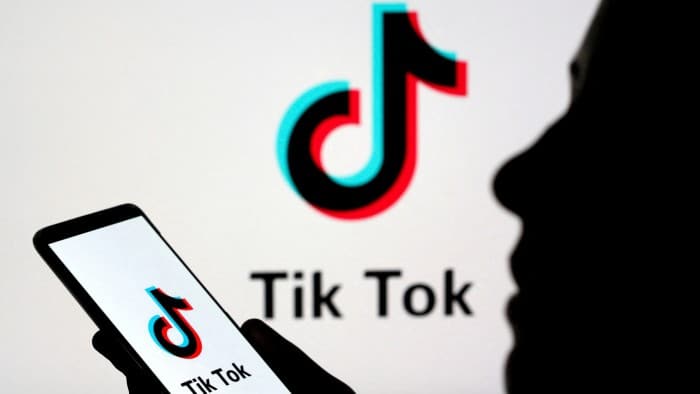 A person holds a smartphone displaying the TikTok logo, with a larger TikTok logo visible in the background