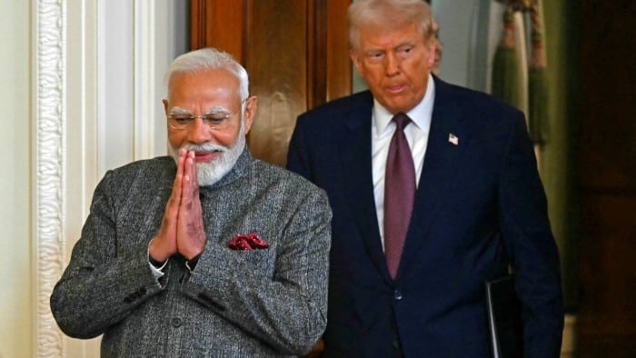 Donald Trump and Narendra Modi walk together in the East Room of the White House. Modi is smiling with his hands in a traditional greeting gesture, while Trump follows behind holding a folder.