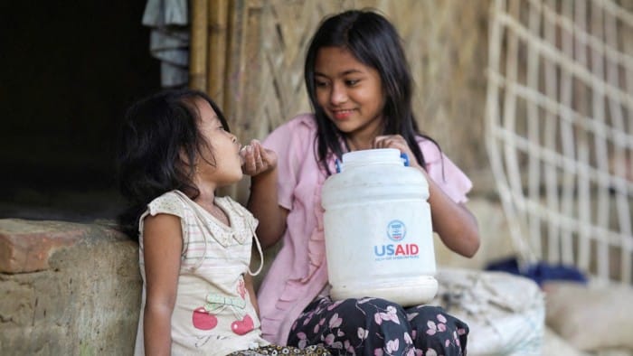 A Rohingya girl feeds a child from a jar with the USAID logo on it