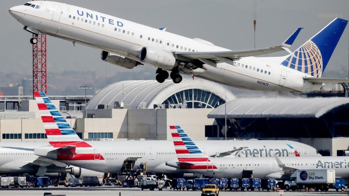 A United Airlines plane is taking off above American Airlines planes parked on the tarmac at Los Angeles International Airport.