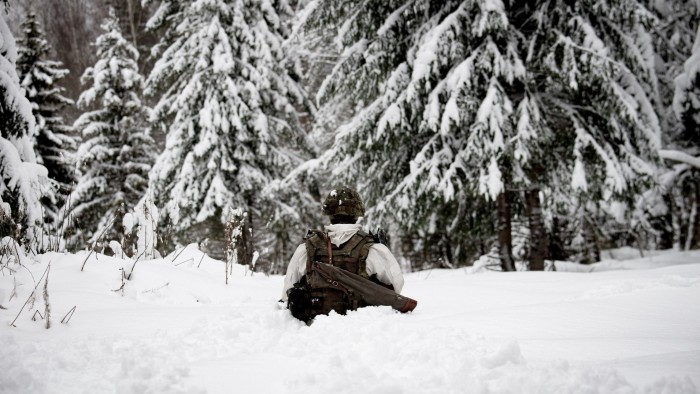 A soldier in winter camouflage gear walks through deep snow during a Nato drill. Snow-covered trees surround the area