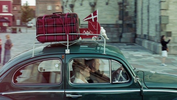 A couple in a green car with red luggage, a small pennant of the Danish flag and a red doll’s pram strapped to the roof, drive through a town