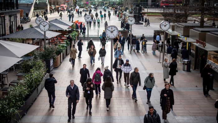 People walk along a bustling pedestrian area in Canary Wharf during lunchtime. The scene is dominated by six large public clocks designed by Konstantin Grcic, which are positioned above the crowd. Cafés and food stalls line the sides, with individuals interacting and queuing.