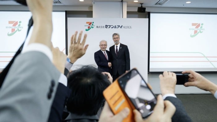 Ryuichi Isaka and Stephen Dacus stand side by side shaking hands in front of a backdrop displaying the company logo while they are surrounded by people taking photos and raising hands during a news conference