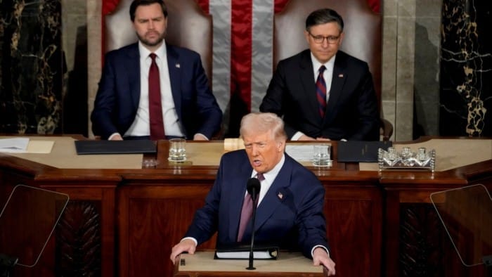 Donald Trump speaks at a lectern with JD Vance and Mike Johnson seated behind him listening attentively