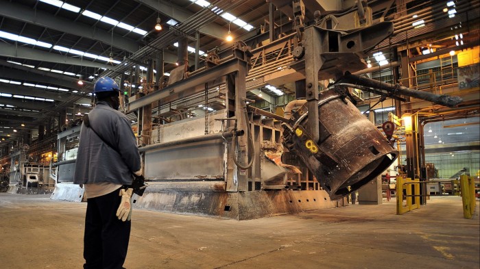 A metal handler stands in a factory wearing a blue helmet and gloves, using a remote control to operate machinery pouring molten aluminium into a furnace