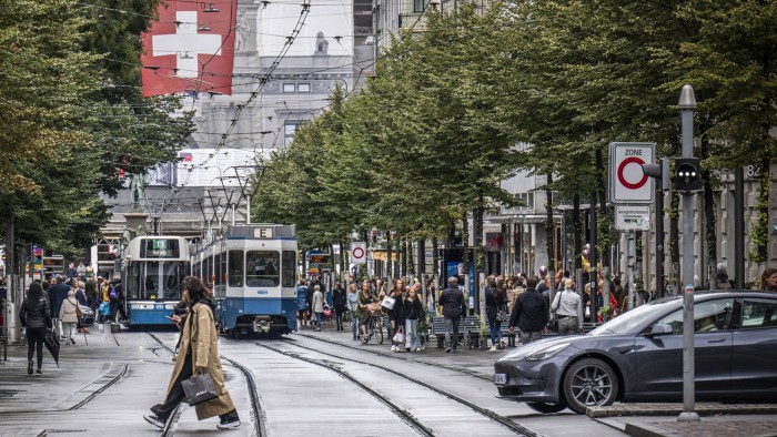 Pedestrians cross tram tracks in a busy shopping area in Zurich. Two trams are visible on the tracks, with numerous people walking along the sidewalks and crossing the street