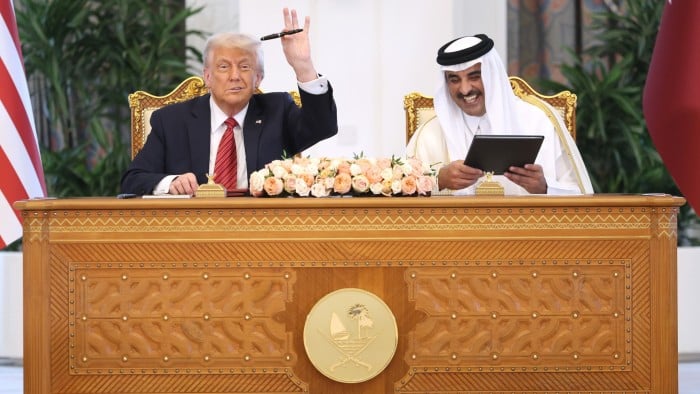 Donald Trump and Tamim bin Hamad al-Thani sit at a desk together signing documents. Trump is gesturing to the camera
