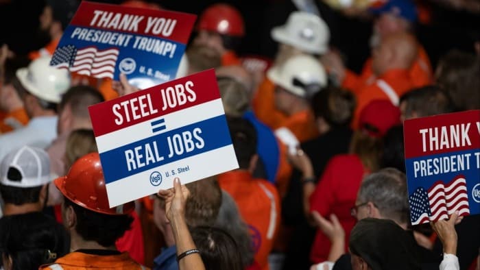 Attendees hold signs reading ‘Steel Jobs = Real Jobs’ and ‘Thank You President Trump!’ with the US Steel logo and American flag motifs. People in the crowd are wearing hard hats and orange work attire