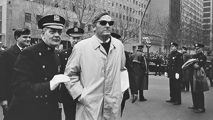 A middle-aged man wearing a light-coloured coat and sunglasses walks with a uniformed police officer. A crowd of protesters is visible in the distance