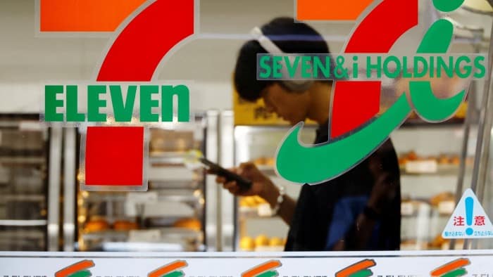 A Tokyo 7 Eleven store, with the logo in the foreground. Seen through the glass is a man checking his phone