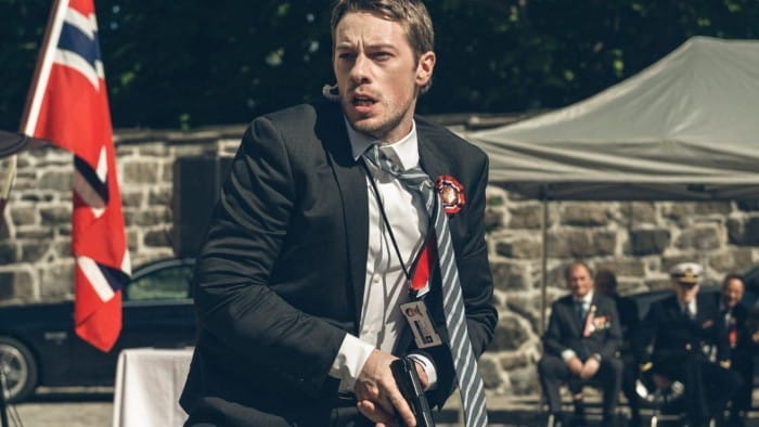 Eldar Skar in a tense scene from ‘Occupied’, wearing a suit and holding a gun, with Norwegian flags and officials in the background