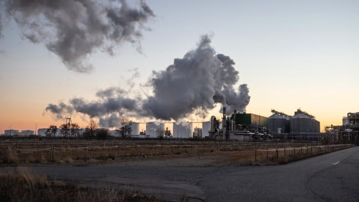 A large refinery complex emitting thick plumes of smoke at sunset, with storage tanks and industrial structures in view.