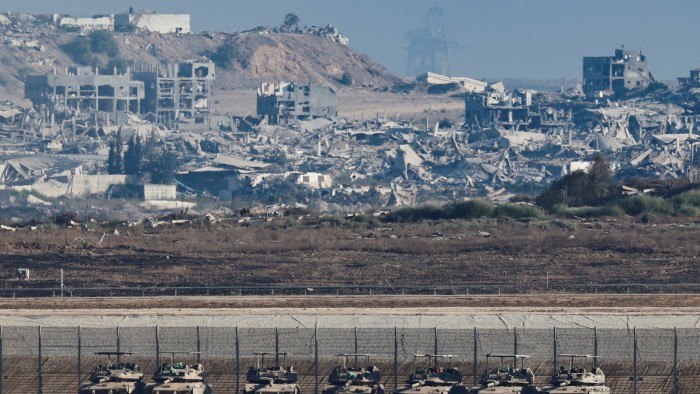 Several tanks are lined up behind a border fence, with widespread destruction and ruined buildings visible in Gaza in the background.